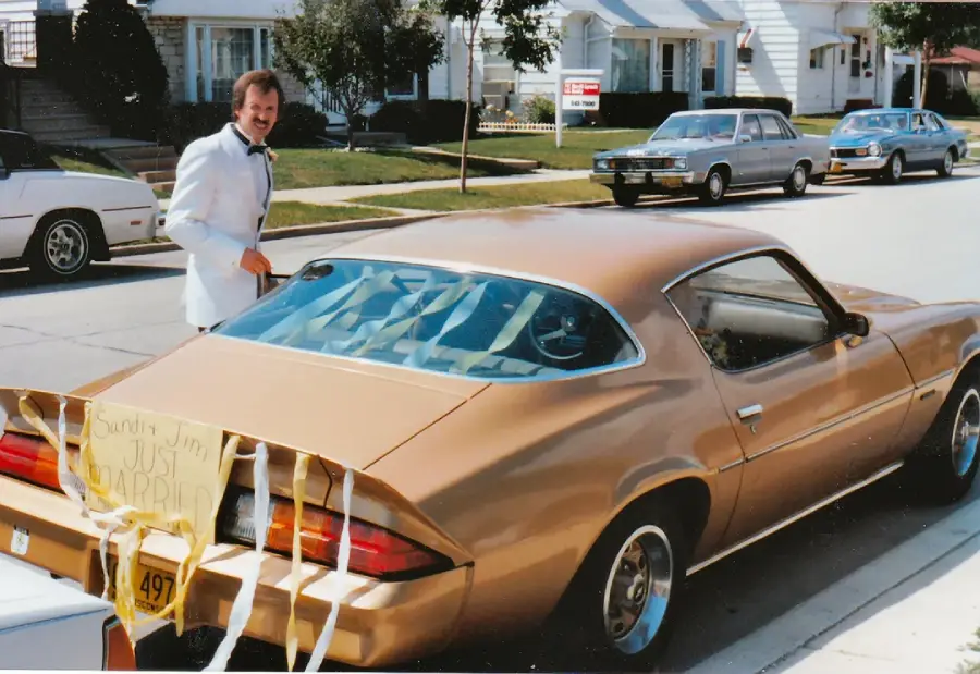 A man in a white suit stands next to a gold car decorated with a "Just Married" sign and streamers, parked on a suburban street lined with houses and other cars.