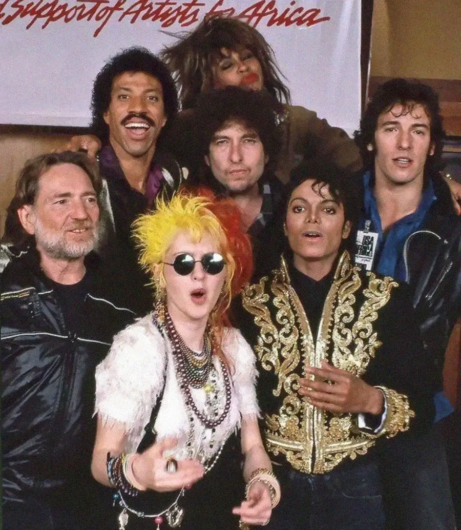 Seven famous musicians from the 1980s, including Lionel Richie, Tina Turner, Bob Dylan, Bruce Springsteen, Willie Nelson, Cyndi Lauper, and Michael Jackson, posing together in front of a "Support of Artists for Africa" banner.