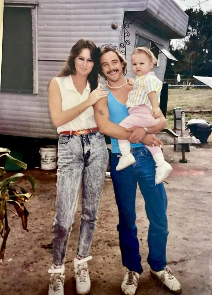A woman and a man holding a small child pose and smile outside in front of a trailer. The woman wears a white sleeveless shirt and jeans, the man wears a tank top and jeans, and the child wears a striped shirt and pink pants.