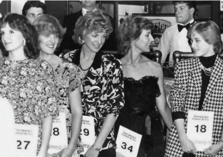 A black-and-white photo shows five women standing in a row at an event, each holding numbered cards. They are dressed in formal attire and appear to be smiling and engaged in conversation. A man in a bow tie stands in the background.