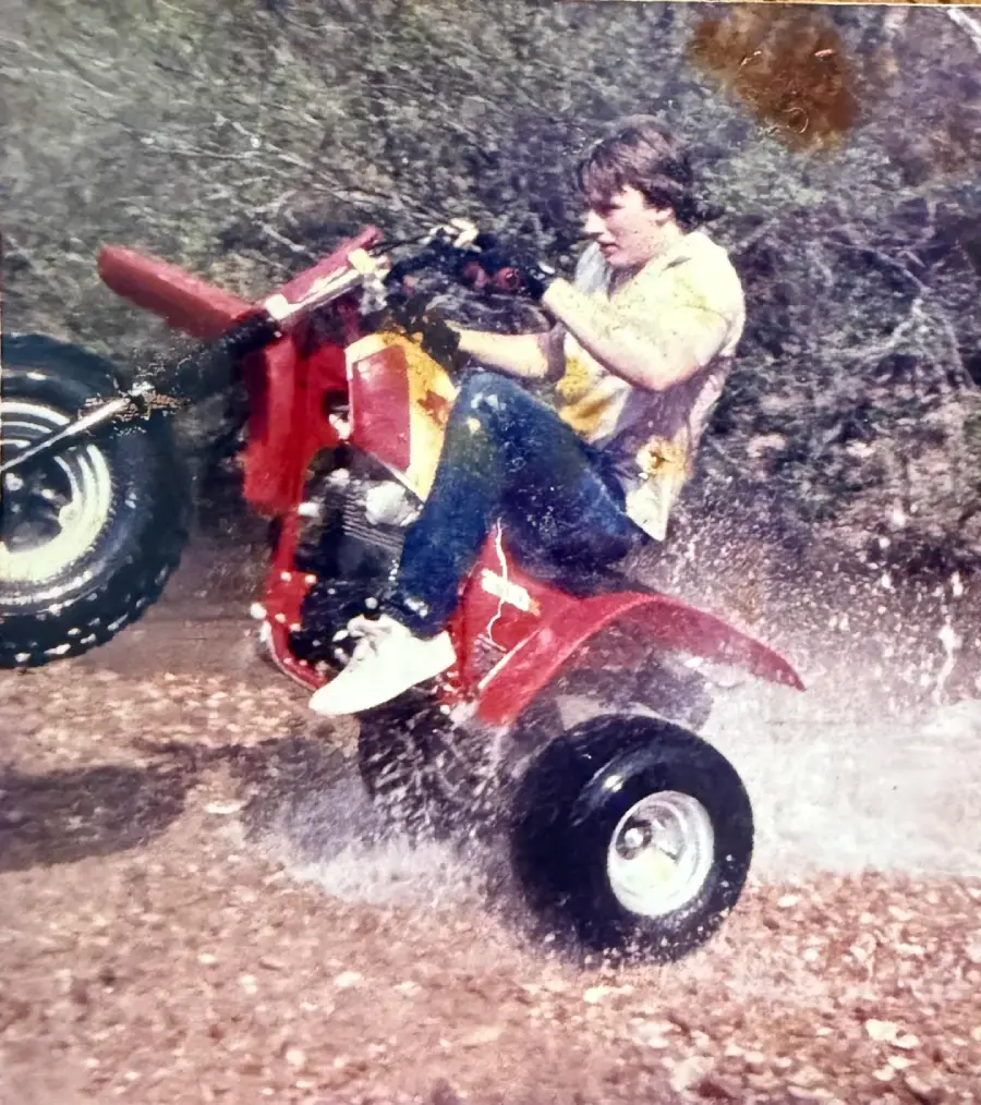 A person wearing jeans and a yellow shirt is riding a red three-wheeler ATV through water, with the front wheels lifted off the ground and water splashing up around them. Trees and foliage are in the background.