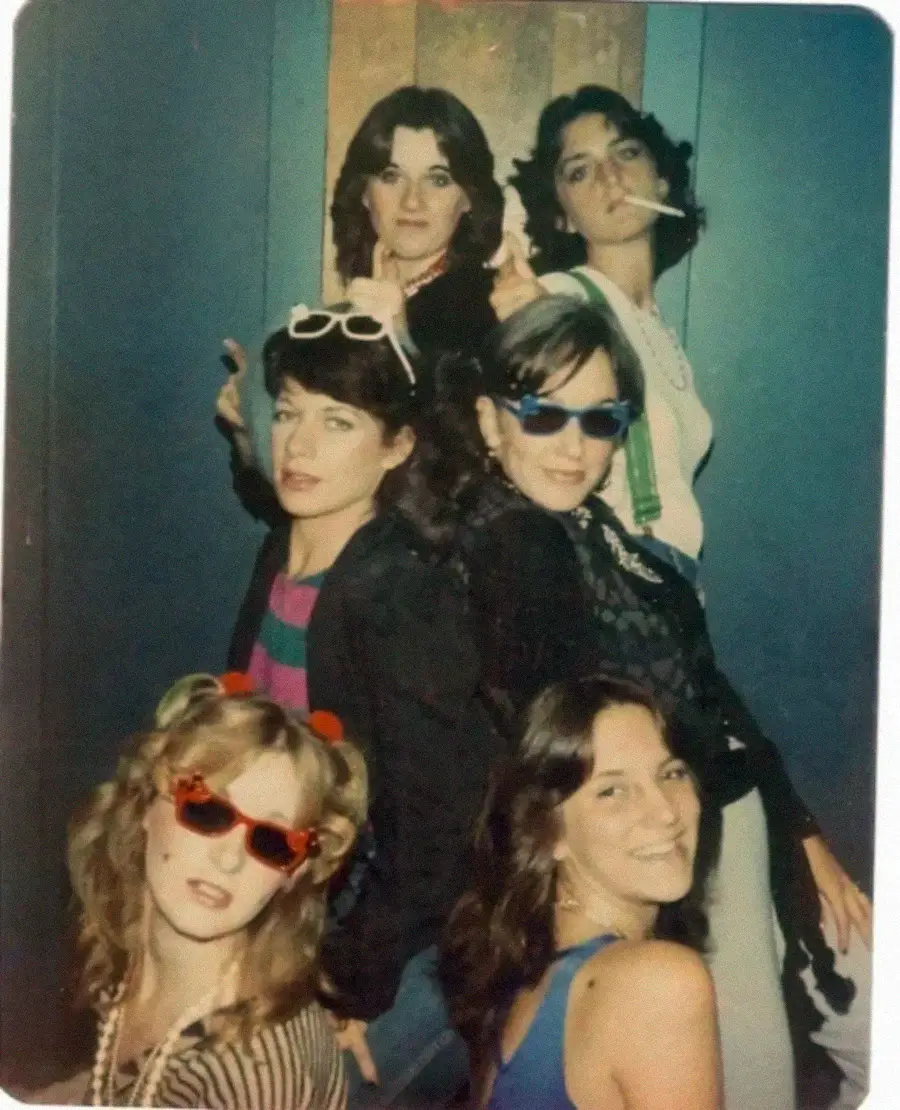 Five women pose playfully in colorful outfits and sunglasses, some with fun accessories including a cigarette and beaded necklaces, in front of a blue wall, evoking a retro, festive vibe.