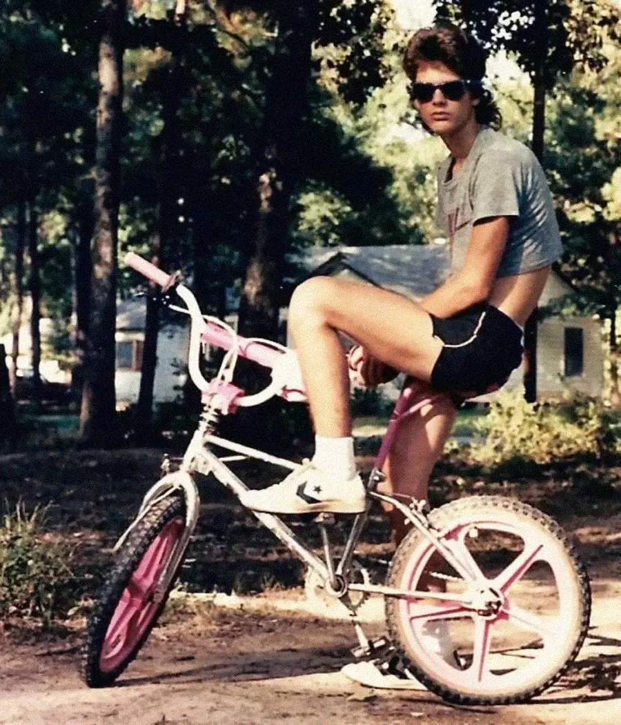 A person wearing sunglasses, a gray crop top, black shorts, and white socks sits sideways on a white and pink BMX bike in a wooded outdoor area with houses in the background.