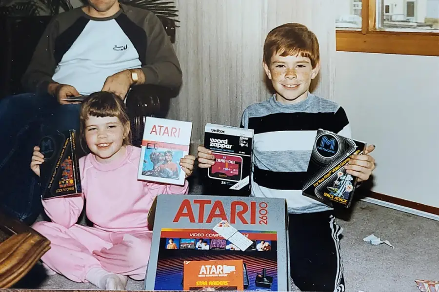 Two smiling children sit on the floor holding Atari video game boxes, with an Atari 2600 console package in front of them. An adult sits behind them, partially visible. They appear happy and excited.