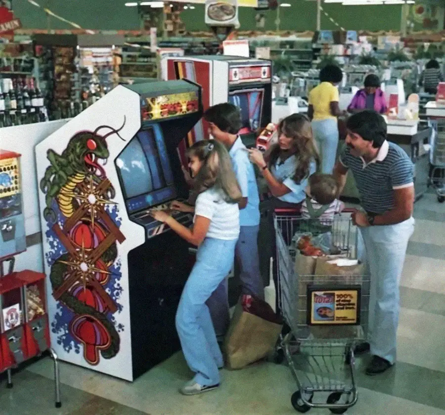 A group of people, including children and adults, play arcade games in a supermarket aisle. Shopping carts and groceries are visible nearby, capturing a retro 1980s atmosphere.