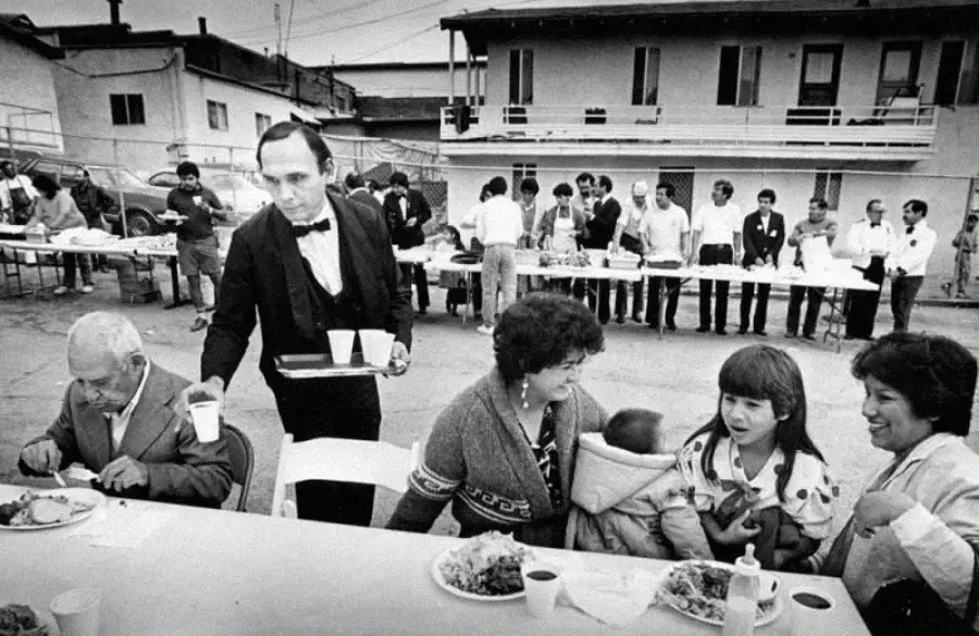 A waiter in a bow tie serves drinks to people seated at a long outdoor table. Behind them, more people gather near a food serving line in a courtyard with buildings in the background.