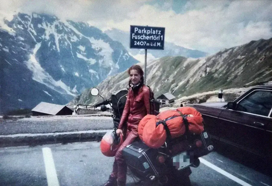 A woman in red motorcycle gear sits on a motorcycle loaded with orange bags in a mountain parking area, with snow-capped peaks in the background and a sign reading "Parkplatz Fuscherörl 2407m ü.d.M.