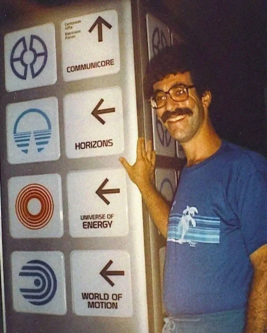 A man with glasses and a mustache smiles while pointing at a sign in a theme park. The sign has arrows and icons for CommuniCore, Horizons, Universe of Energy, and World of Motion attractions.