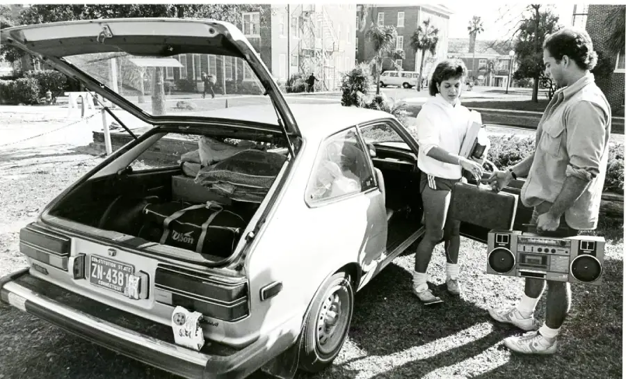 Two people stand near an open hatchback car packed with luggage. One hands something to the other, who holds a large boombox. Buildings, trees, and parked cars are visible in the background on a sunny day.