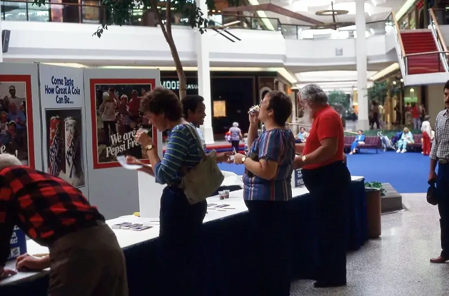 People stand at an information booth in a shopping mall, reading pamphlets and talking. Posters and promotional materials are displayed on the booth. Shoppers and children are visible in the background.
