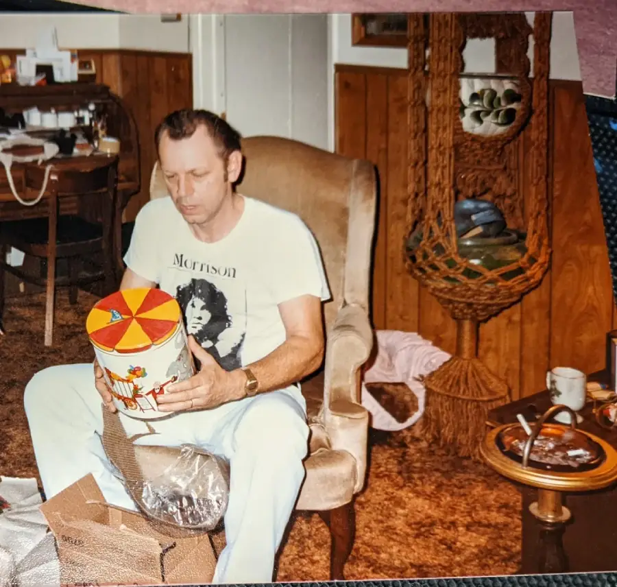 A man in a white "Morrison" T-shirt sits on a beige chair, holding a colorful tin. The room has wood-paneled walls, a table with dishes, and a hanging macramé shelf with bowls. A cup and saucer are on a small side table nearby.