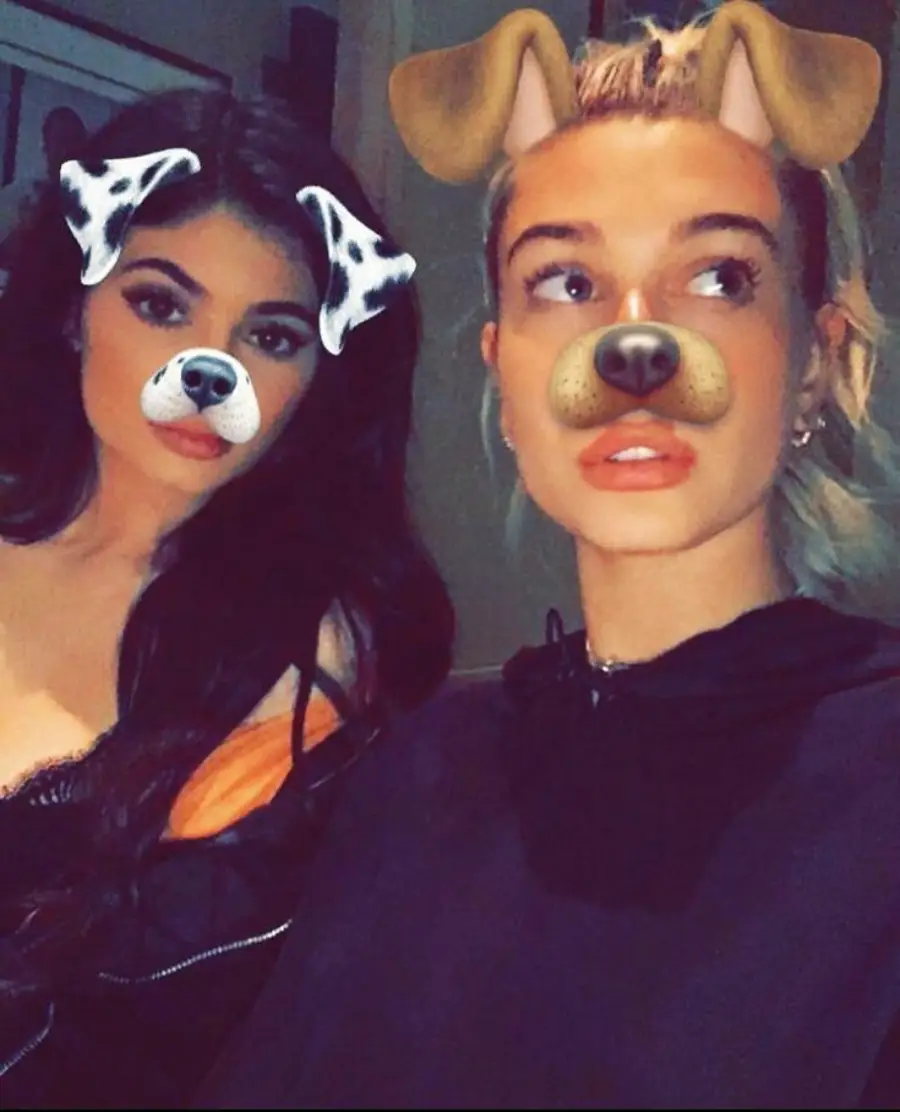 Two women pose for a selfie, both using a dog filter that adds cartoon dog ears and noses to their faces. The woman on the left has long dark hair; the woman on the right has blonde hair pulled back.
