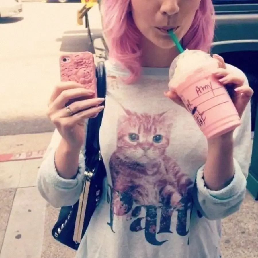 A person with pastel pink hair sips a pink drink through a straw and holds a pink phone. They wear a sweatshirt featuring a large cat graphic and the word “Paris.” The name “Amy” is written on the drink cup.