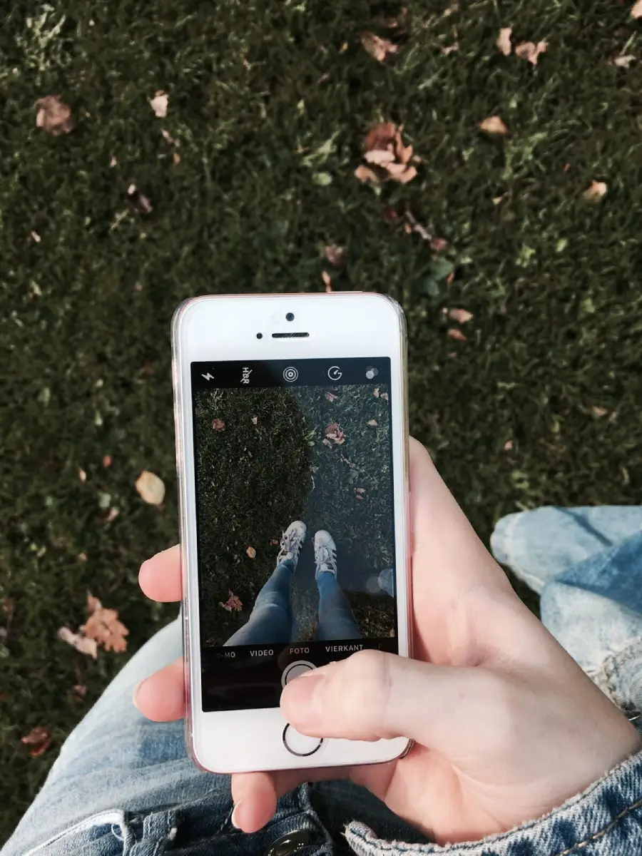 A person holds a smartphone and takes a photo of their feet on grass with scattered leaves. The phone screen shows the camera view of the shoes and ground.
