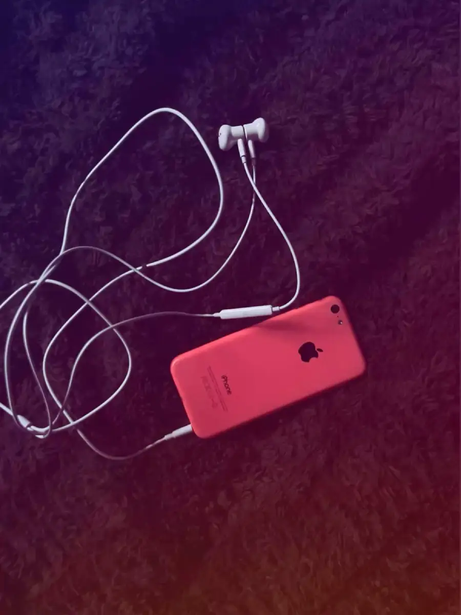 A pink iPhone with white earphones plugged in rests on a dark, textured surface. The earphone cable is loosely coiled around the phone.