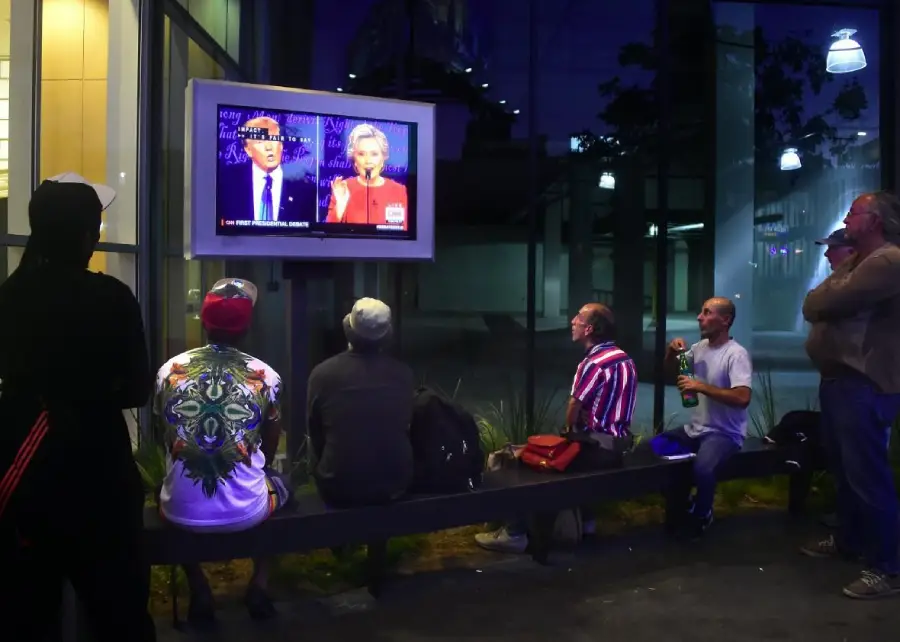 A group of people sit and stand at a bus stop at night, watching a televised debate between two politicians on a screen mounted to the wall. The scene is illuminated by city lights behind glass panels.