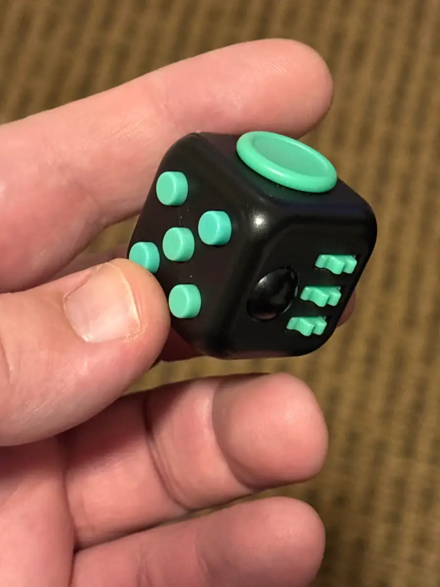 A hand holding a black fidget cube with green buttons, a green dial, and other tactile features. The background is a brown, textured surface.