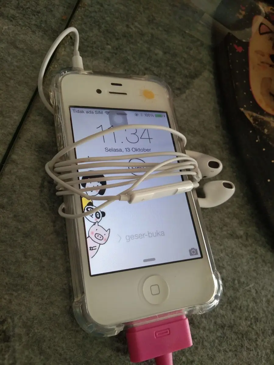 A white iPhone with a cartoon cow wallpaper is lying on a gray surface. White earphones are neatly wrapped around the phone, which is charging with a pink cable. The screen displays the time and date.