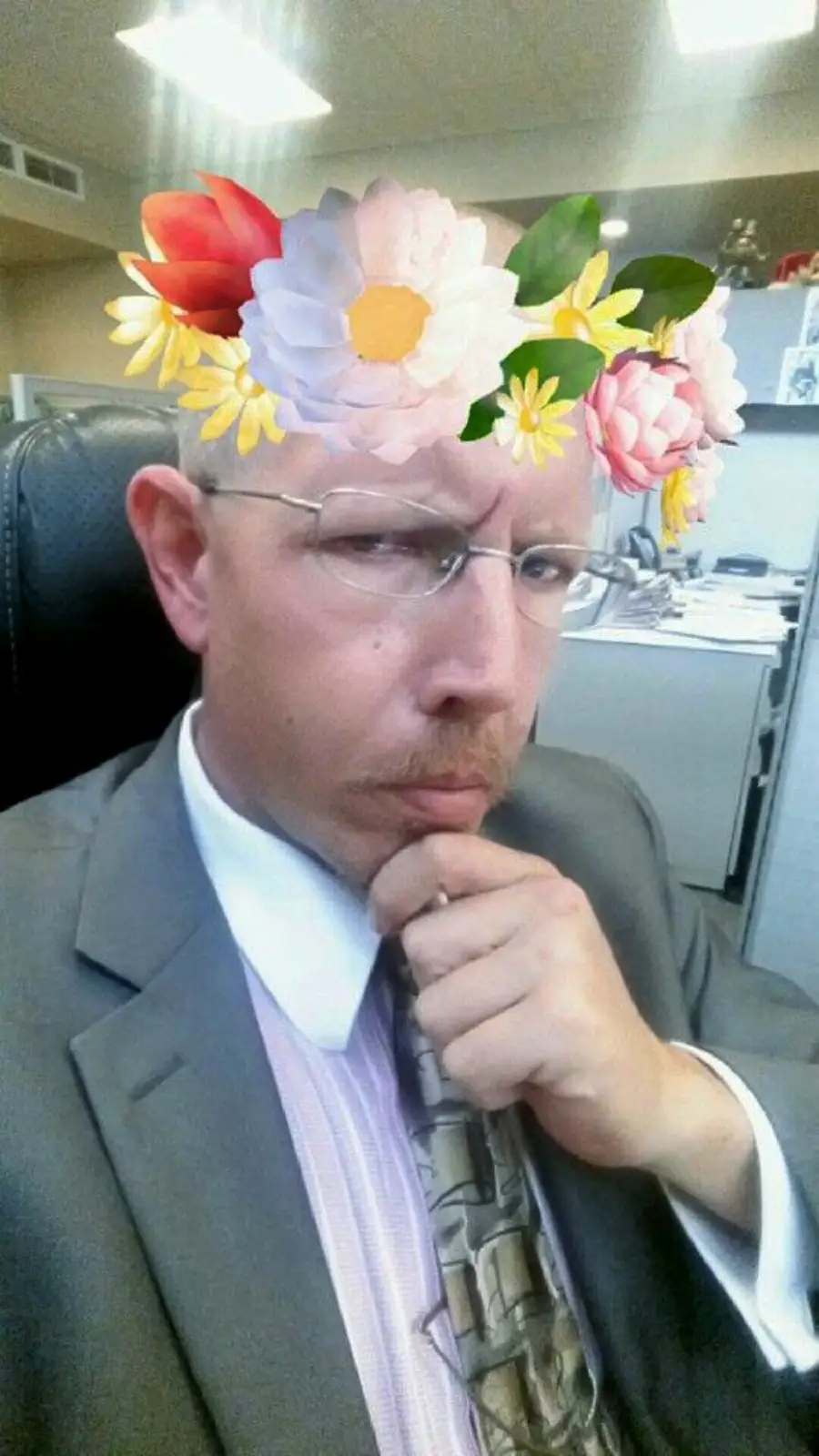 A man in a suit and glasses sits in an office, thoughtfully resting his chin on his hand. He is wearing a playful virtual flower crown filter on his head.