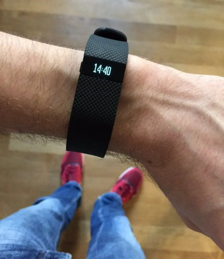 A person wearing a black fitness tracker displaying the time 14:40 stands on a wooden floor, wearing blue jeans and red sneakers.