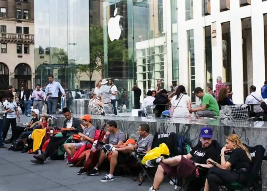 People sit and wait in a long line outside a large Apple Store with a glass facade in an urban area, some lounging in chairs and using electronic devices. The Apple logo is prominently displayed on the building.