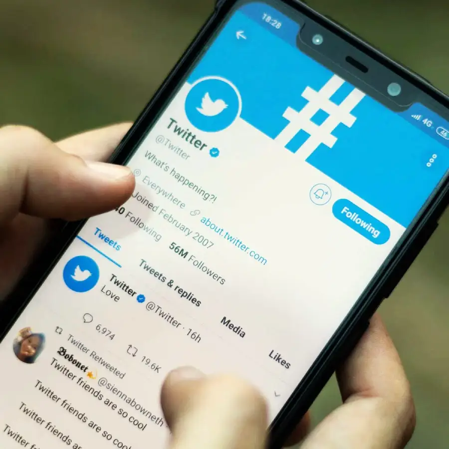 A person holds a smartphone displaying the Twitter profile page, showing the Twitter logo, account info, and blue interface, with a finger poised above the screen.