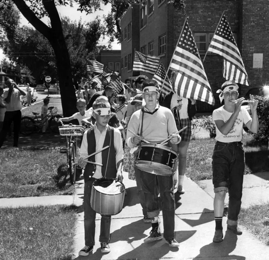 A group of children march on a sidewalk, carrying American flags and playing drums and a flute. The scene appears festive, with more people and flags in the background on a sunny day.