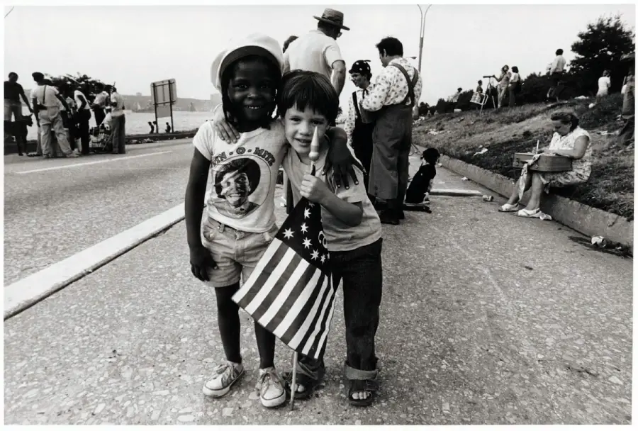 Two young children, one Black and one white, stand on a street smiling with their arms around each other. They hold an American flag, with people gathered in the background.