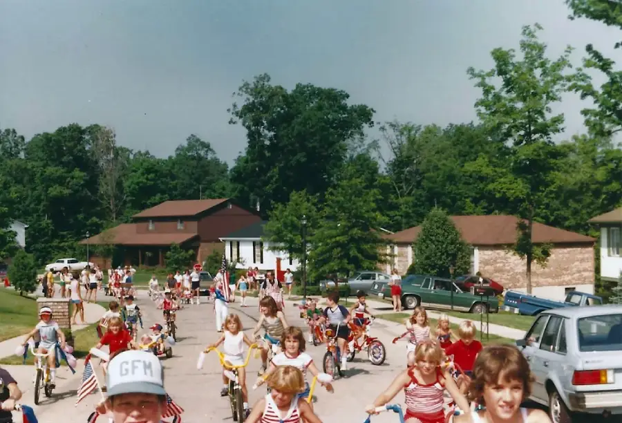 Children ride decorated bicycles down a suburban street, many wearing red, white, and blue outfits. Houses, trees, and parked cars line the road, suggesting a festive parade, possibly for the Fourth of July.