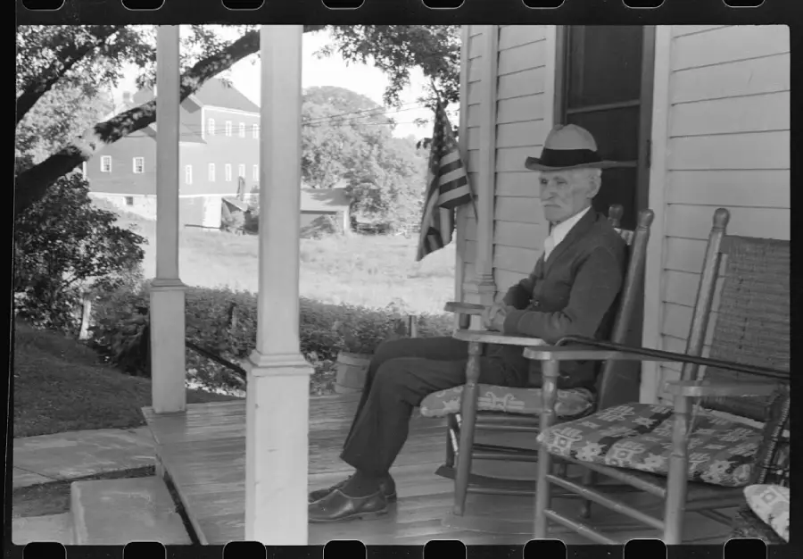 An older man in a suit and hat sits on a rocking chair on a porch, arms crossed. An American flag hangs nearby, trees and a large building are visible in the background.