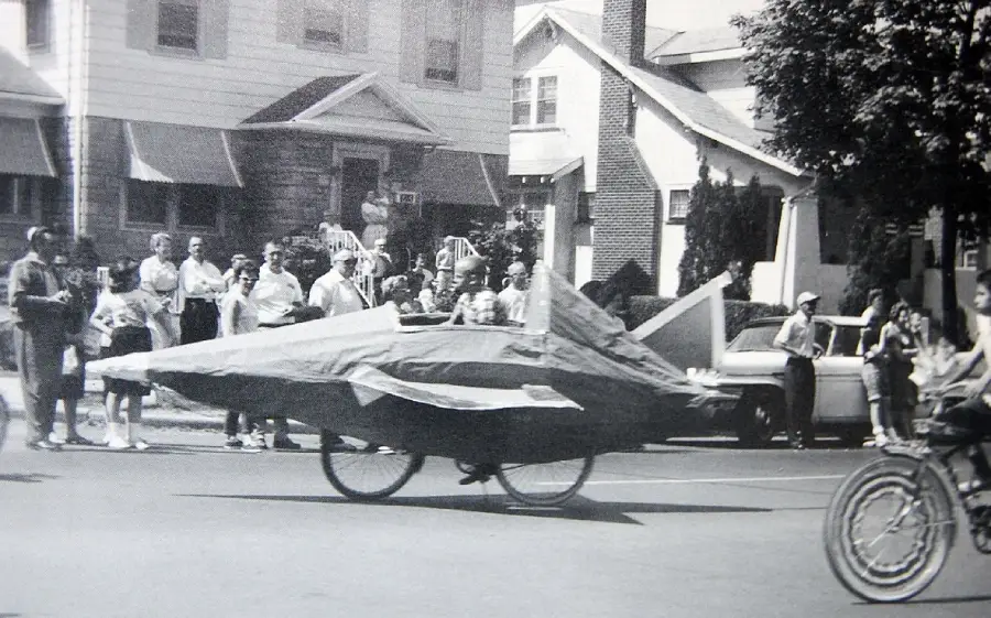 Black and white photo of a parade, showing a person riding a bicycle covered with a homemade rocket ship costume. A crowd of people watches from the sidewalk in front of houses, and another person rides a bicycle nearby.