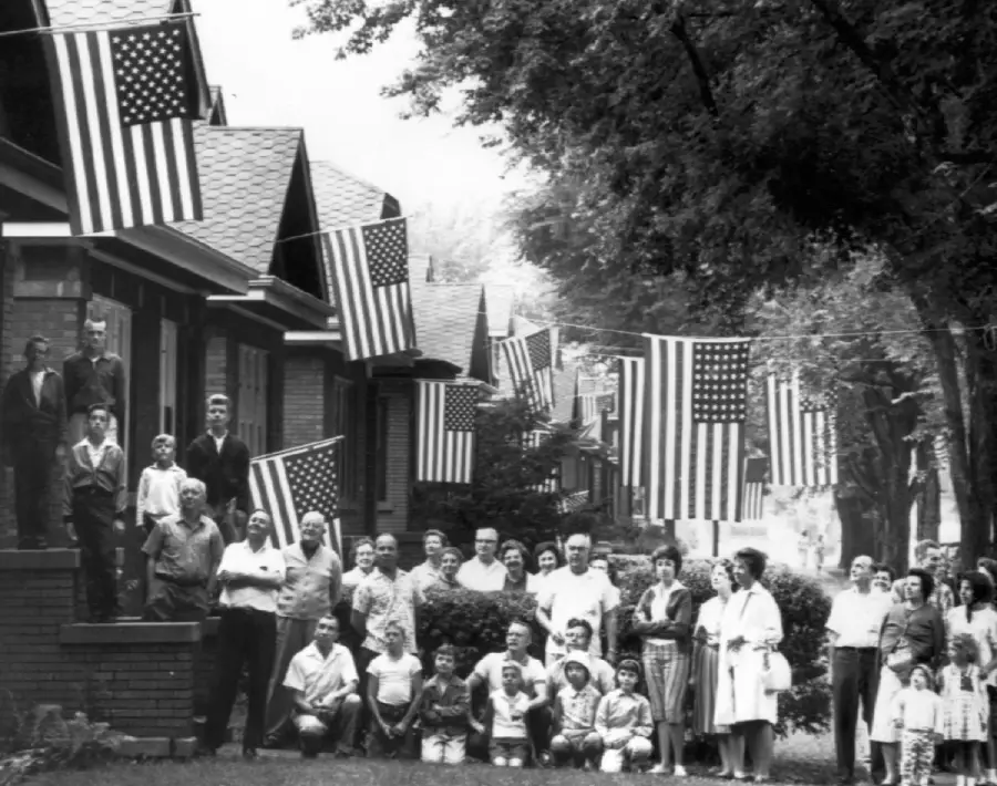 A large group of people, including children and adults, pose on a residential street lined with American flags hung from houses. The scene appears to be from a past era, suggesting a patriotic celebration.