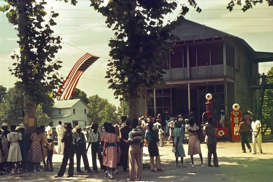 A group of people, mostly dressed in 1940s clothing, gather outside a building with two vintage gas pumps and a large American flag hanging above on a sunny day. Tall trees partially shade the crowd.