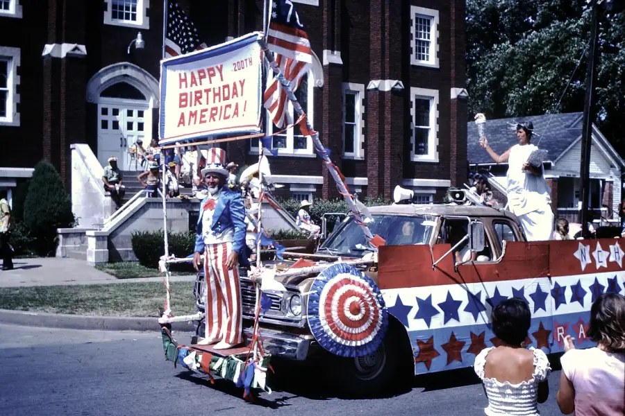 A parade float decorated with American flags, stars, and red, white, and blue colors carries people in patriotic costumes and a “Happy Birthday America!” sign in front of a brick building. Spectators watch from the street.
