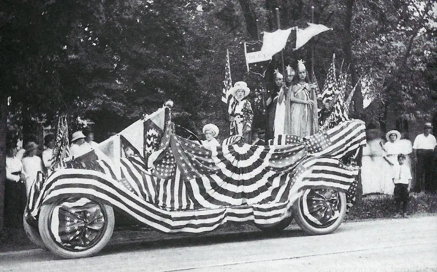A vintage parade car covered in American flags and bunting, with costumed people standing on it holding flags. Spectators dressed in early 1900s clothing watch from the sidewalk under trees.