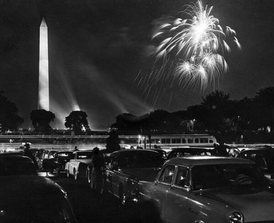 Black-and-white photo of cars parked near the Washington Monument at night, with fireworks lighting up the sky and spotlights shining from the monument, creating a festive atmosphere.