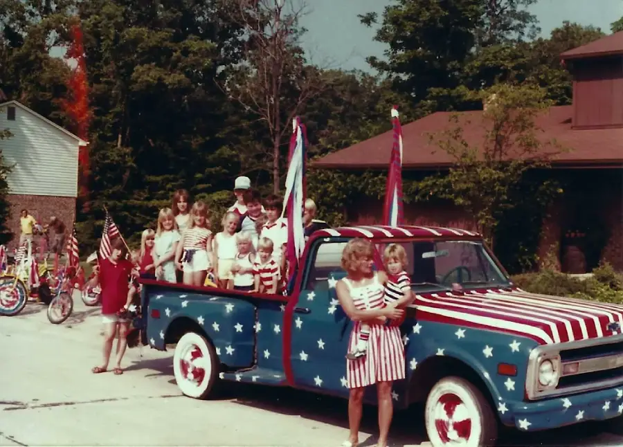 A group of children and adults, some holding American flags, stand by and in the bed of a vintage pickup truck painted with stars and stripes. The scene appears festive, with bicycles and houses in the background.