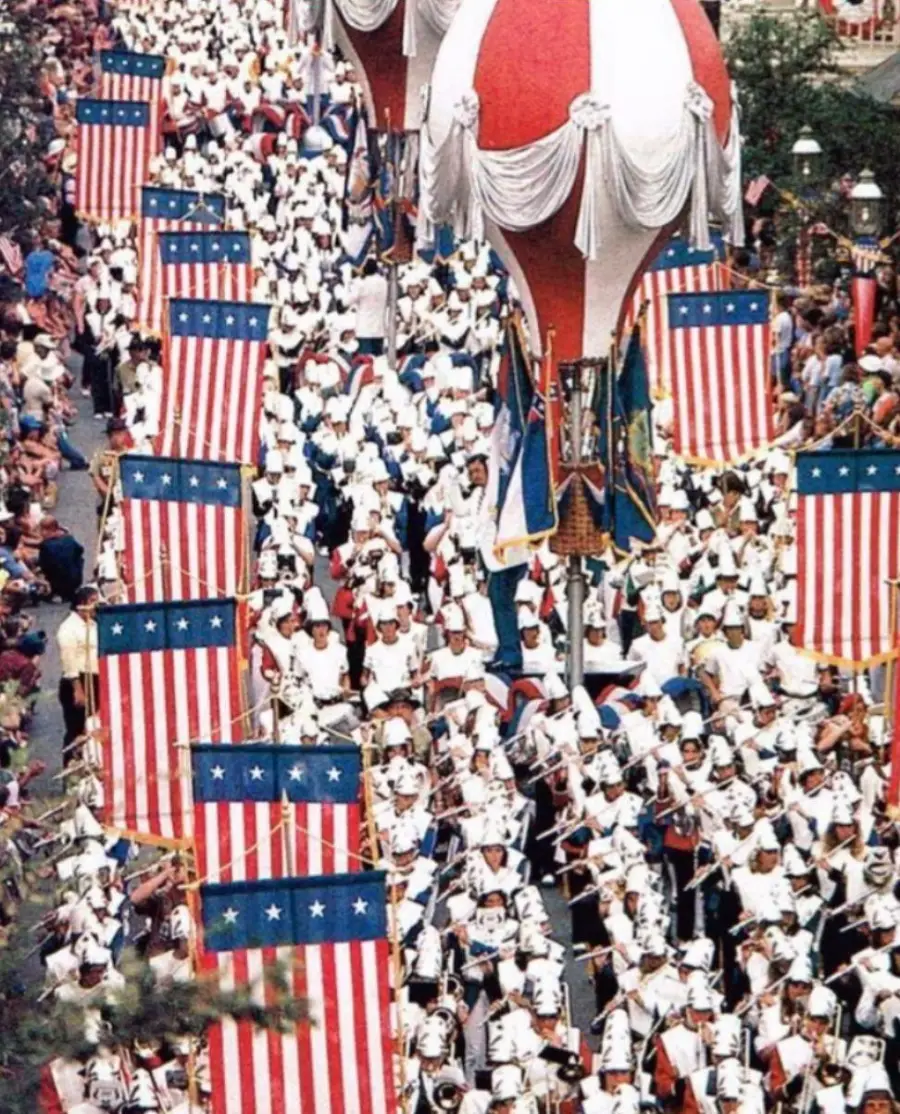 A large marching band in white uniforms parades down a street lined with crowds, carrying American flags and playing instruments, with big red, white, and blue hot air balloon decorations overhead.