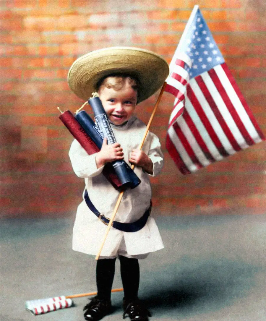 A young child in a straw hat and white outfit holds large mock firecrackers and an American flag, standing in front of a brick wall. The child smiles at the camera, evoking a festive patriotic theme.