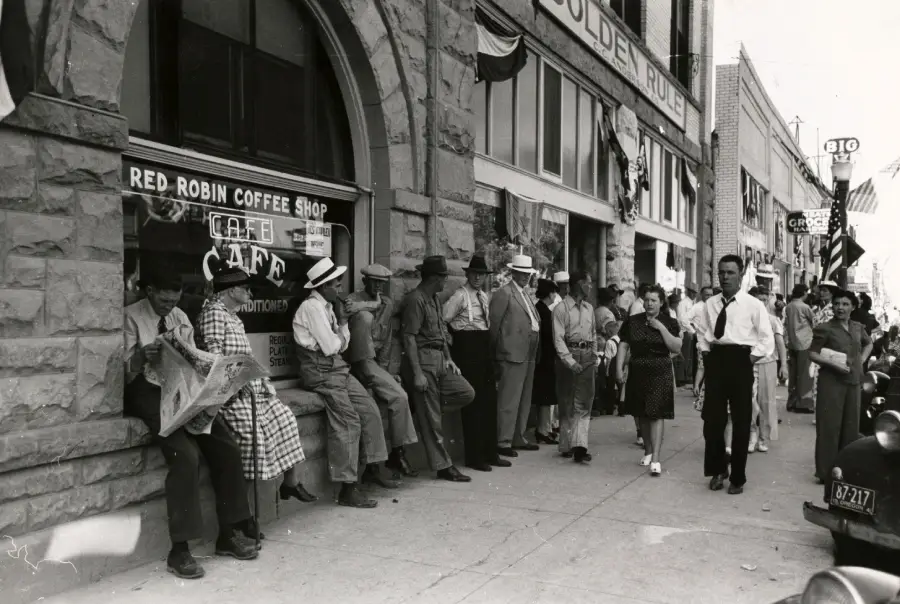 Black-and-white photo of people standing and sitting outside a coffee shop on a city street. Some read newspapers, others talk; storefront signs and an American flag are visible. The scene appears to be from the early-to-mid 20th century.