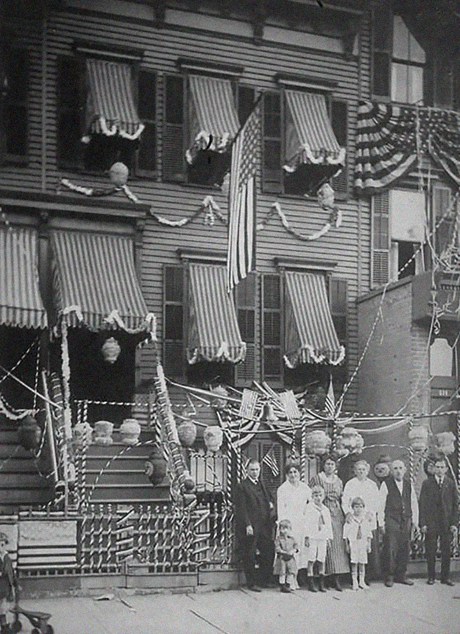 A black-and-white photo of a group of people, including children and adults, standing in front of a decorated house with striped awnings, bunting, and a large American flag. The house is adorned for a celebration.
