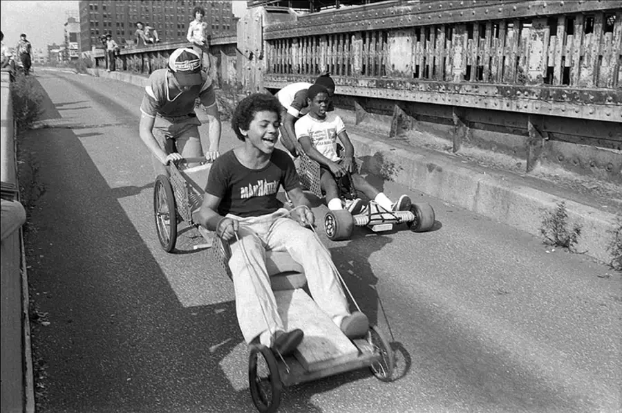 Three kids race homemade go-karts down a city street, smiling and laughing. The scene is energetic, with urban buildings and a metal railing in the background.