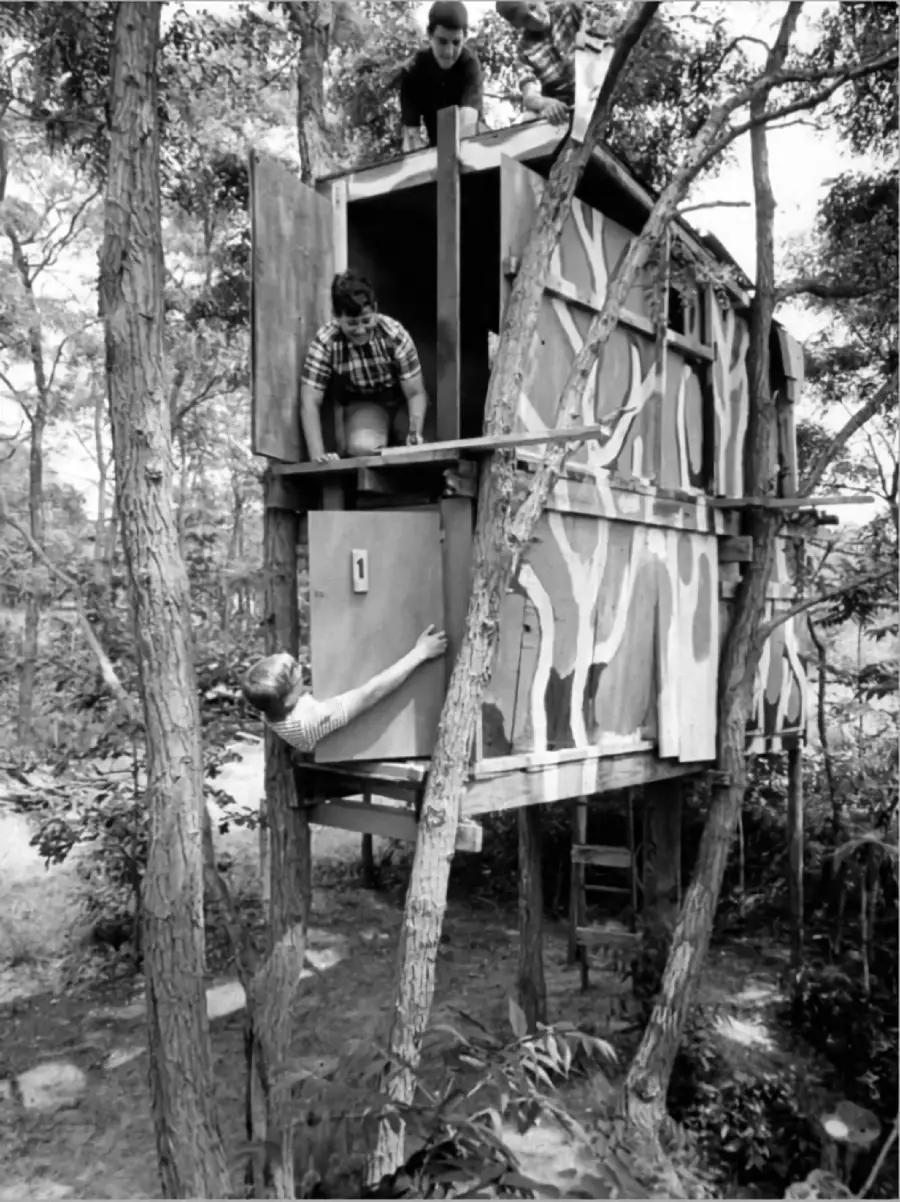 Three boys work together on a two-story treehouse in a wooded area. One boy is on top, another leans out of an upper door, and a third hands up a door from below. The treehouse is painted with abstract patterns.