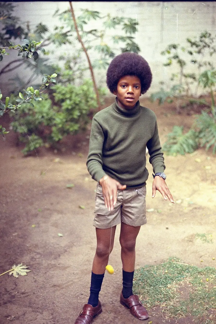 A young boy with an afro hairstyle stands outdoors on dirt and grass, wearing a green turtleneck sweater, khaki shorts, long dark socks, and brown shoes, with a yellow yo-yo dangling from his hand. Shrubs and plants are in the background.