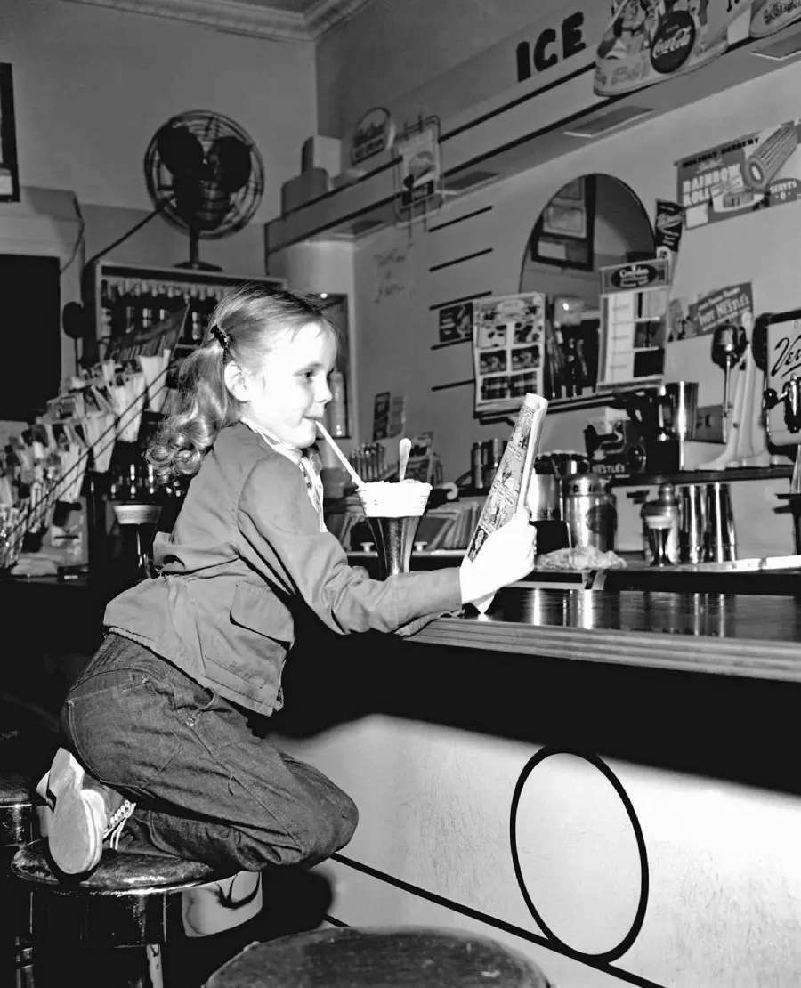 A young girl kneels on a stool at a soda fountain counter, sipping a drink through a straw and reading a comic book. The background features drink machines, posters, and various decorations.