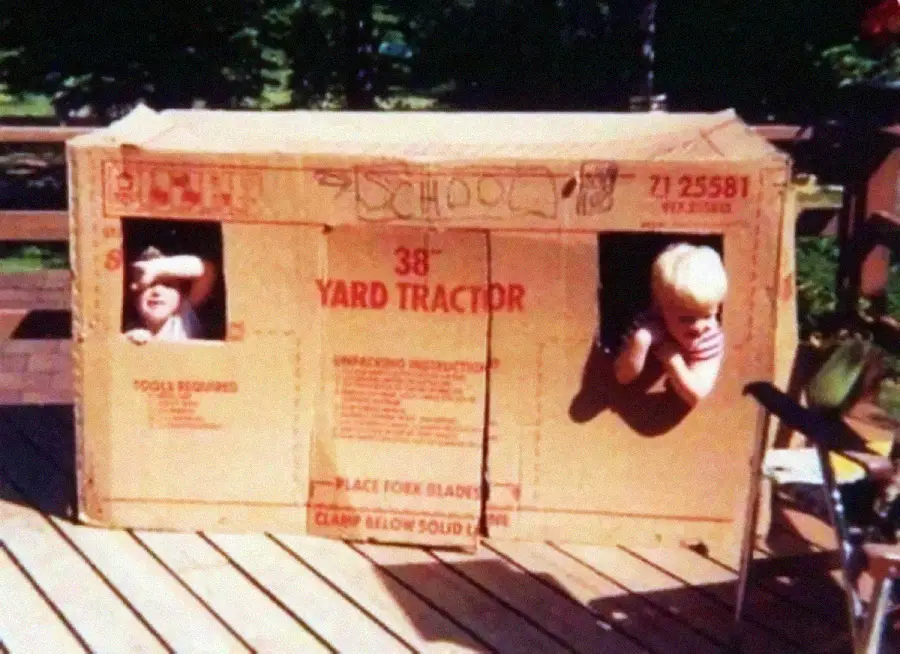 Two children peek out of window cutouts in a large cardboard box labeled "38'' Yard Tractor," sitting on a wooden deck surrounded by outdoor greenery.