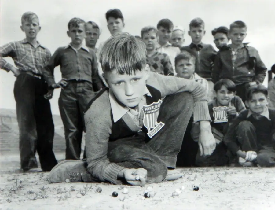 A group of boys watch as a boy kneels on the ground, focused on playing marbles. The other boys stand or crouch behind him, all outdoors on a dirt patch. The image is black and white and appears vintage.