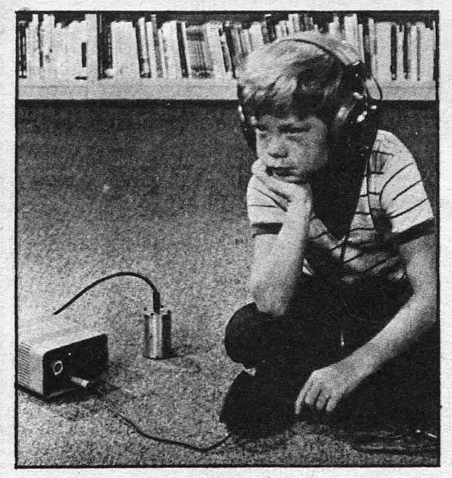 A boy wearing headphones sits on the floor, looking thoughtful, with his elbow resting on his knee. He is connected to a small electronic device placed on the carpet. A bookshelf is visible in the background.
