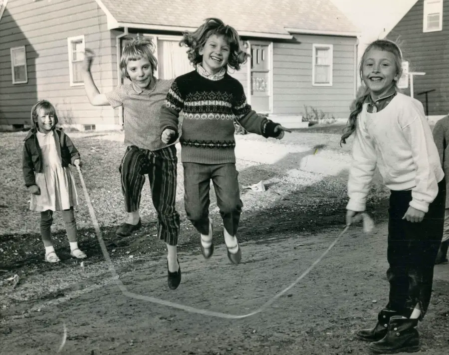 Four young girls play jump rope on a dirt road in front of houses. Two girls jump in the middle, one turns the rope, and another stands nearby watching. All are smiling and dressed in casual clothes. The scene appears vintage.