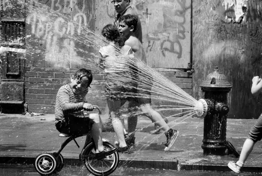 Three children play joyfully around an open fire hydrant spraying water on a city sidewalk. One child rides a tricycle, while two others run barefoot, laughing as water splashes over them. Graffiti covers the wall behind them.